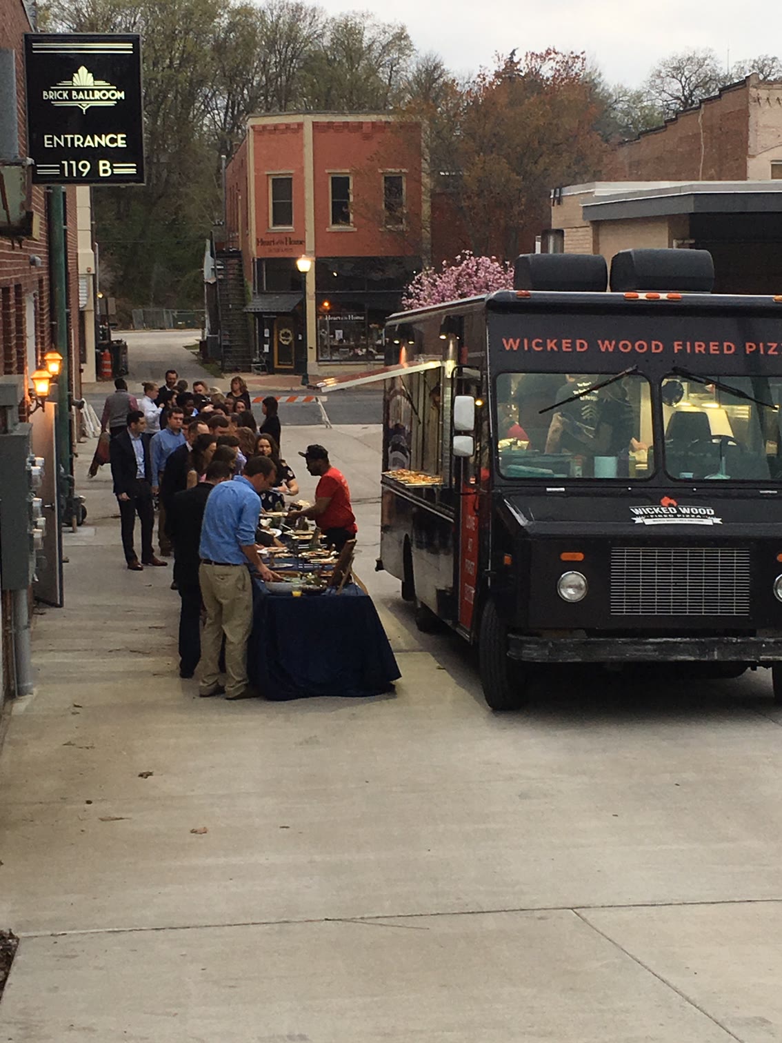 Guests lined along a table in the alley beside the building, a wood-fired pizza truck parked alongside and the Brick Ballroom entrance sign overhead.