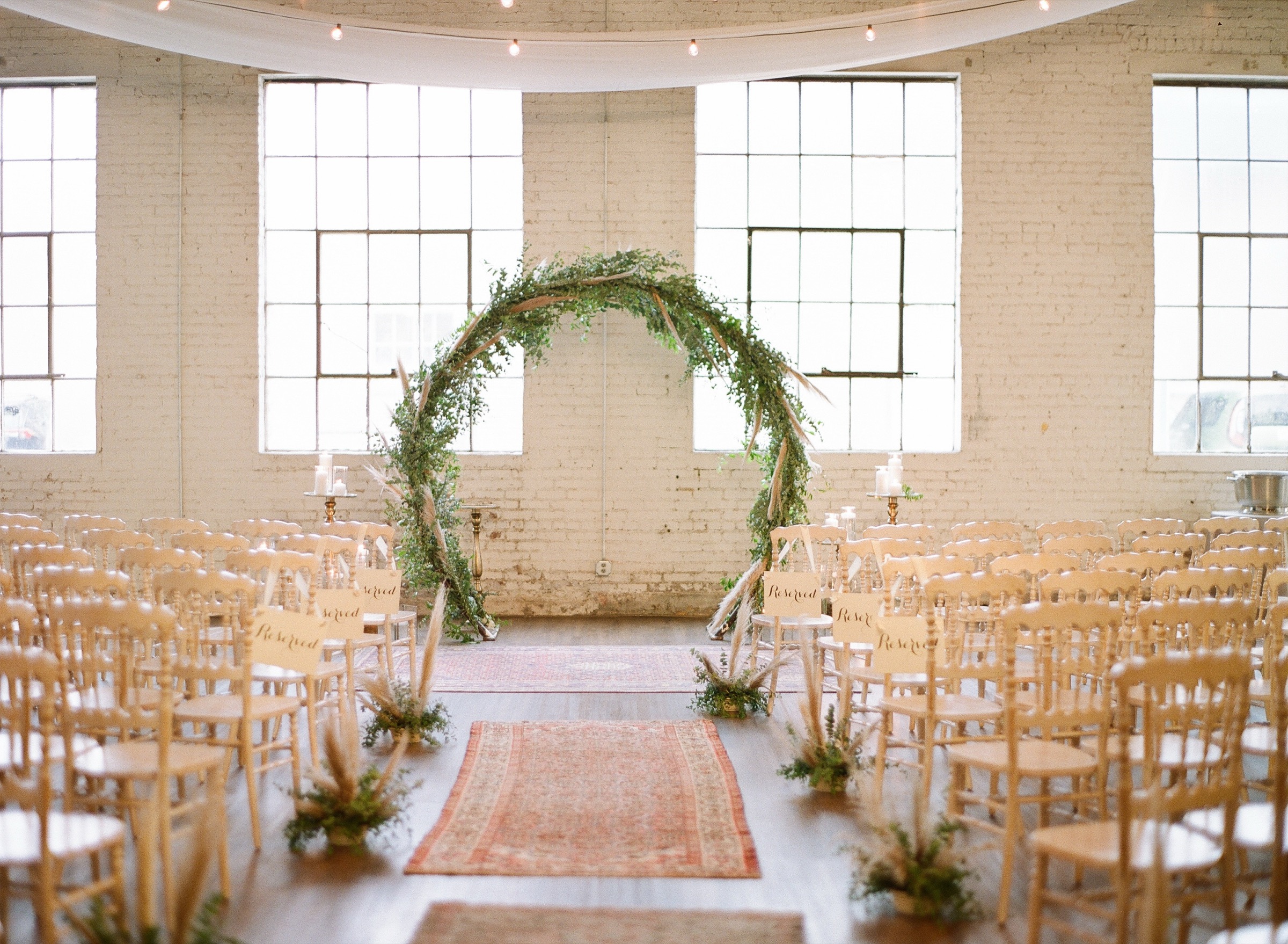The ballroom set and waiting for a ceremony: a circular greenery arch between tall steel windows, chairs in rows on the aisle runner.