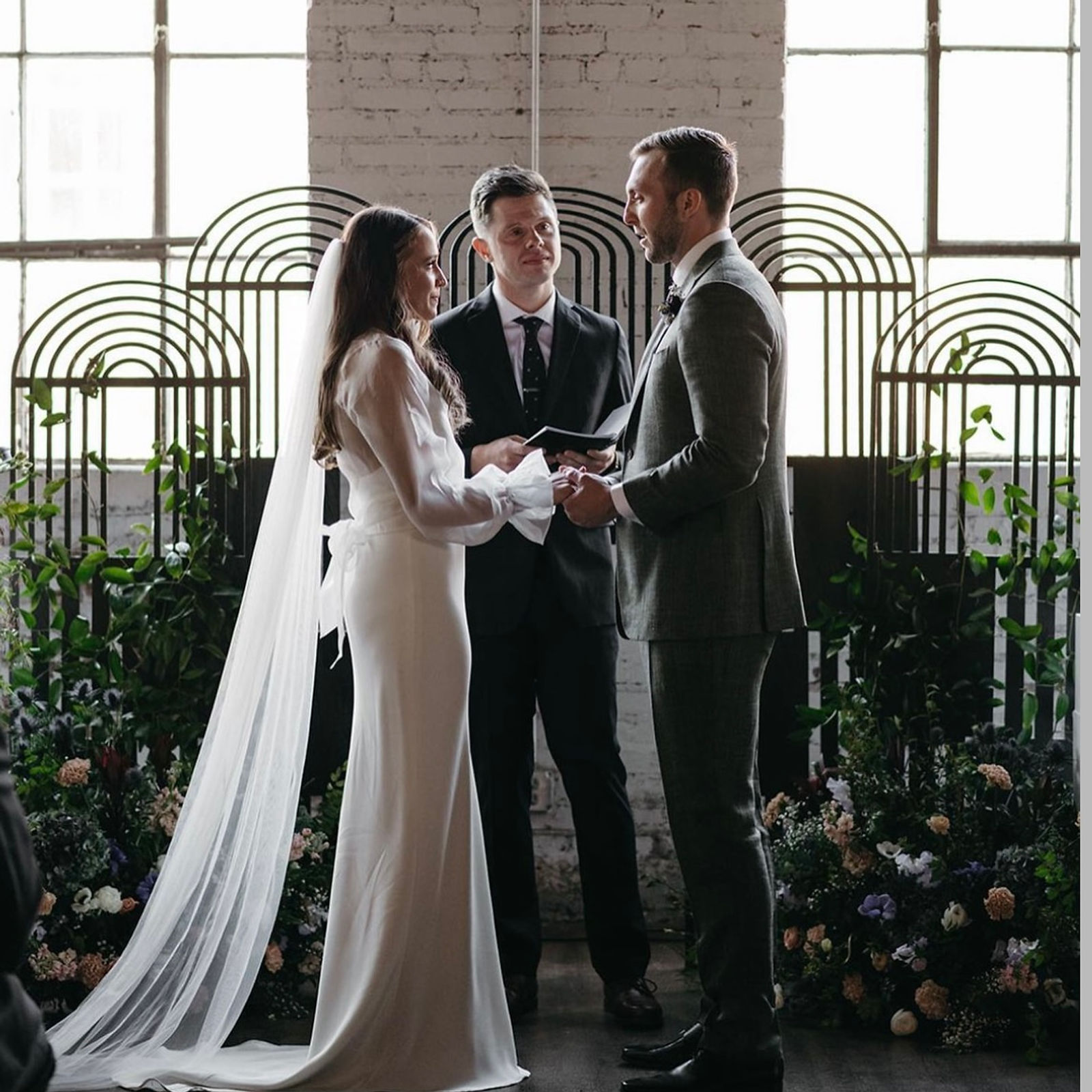 A couple exchanging vows in front of a black arched steel screen, backlit by the ballroom's windows.