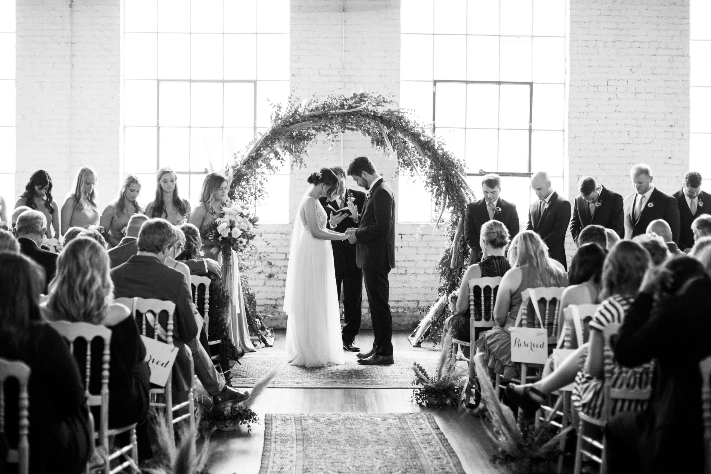 A full congregation seated for a ceremony beneath a circular greenery arch, tall steel windows behind.