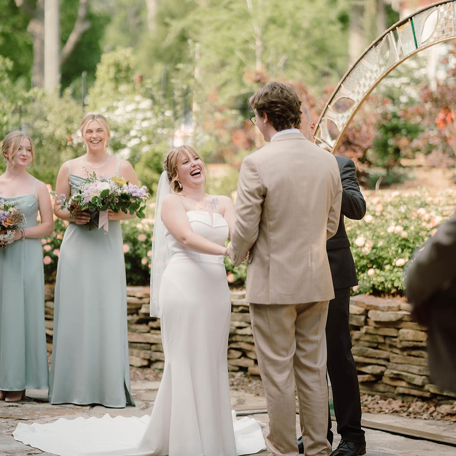 An outdoor garden ceremony — vows beneath a mosaic arch, roses and a stacked-stone wall behind.