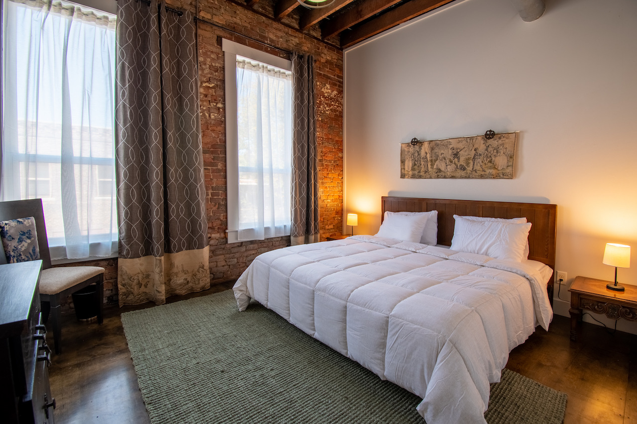 A second bedroom, warm lamps at either side of a wood-panelled headboard, brick along one wall and curtained windows along the other.