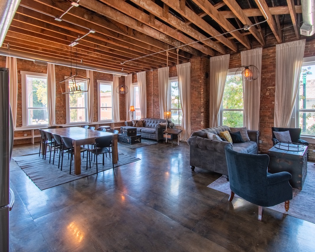 The open living and dining room: exposed wood joists, red brick walls, tall curtained windows over downtown, tufted grey sofas and a long wood table on polished concrete.