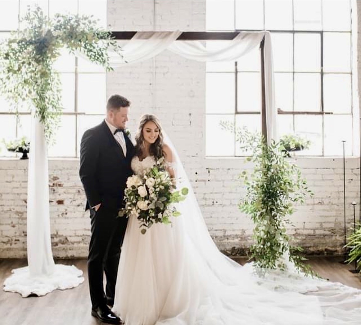 The square arbor dressed with white drapery and greenery, a couple standing beneath it in front of the ballroom's tall windows.
