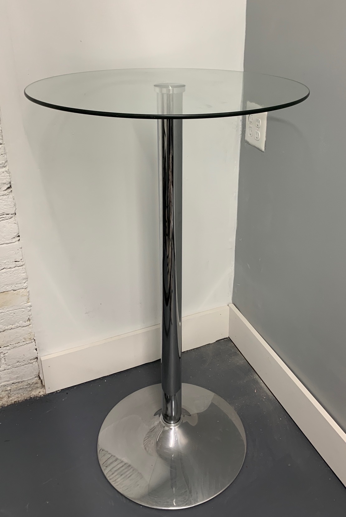 A bar-height round glass cocktail table on a slim chrome pedestal with a weighted base.
