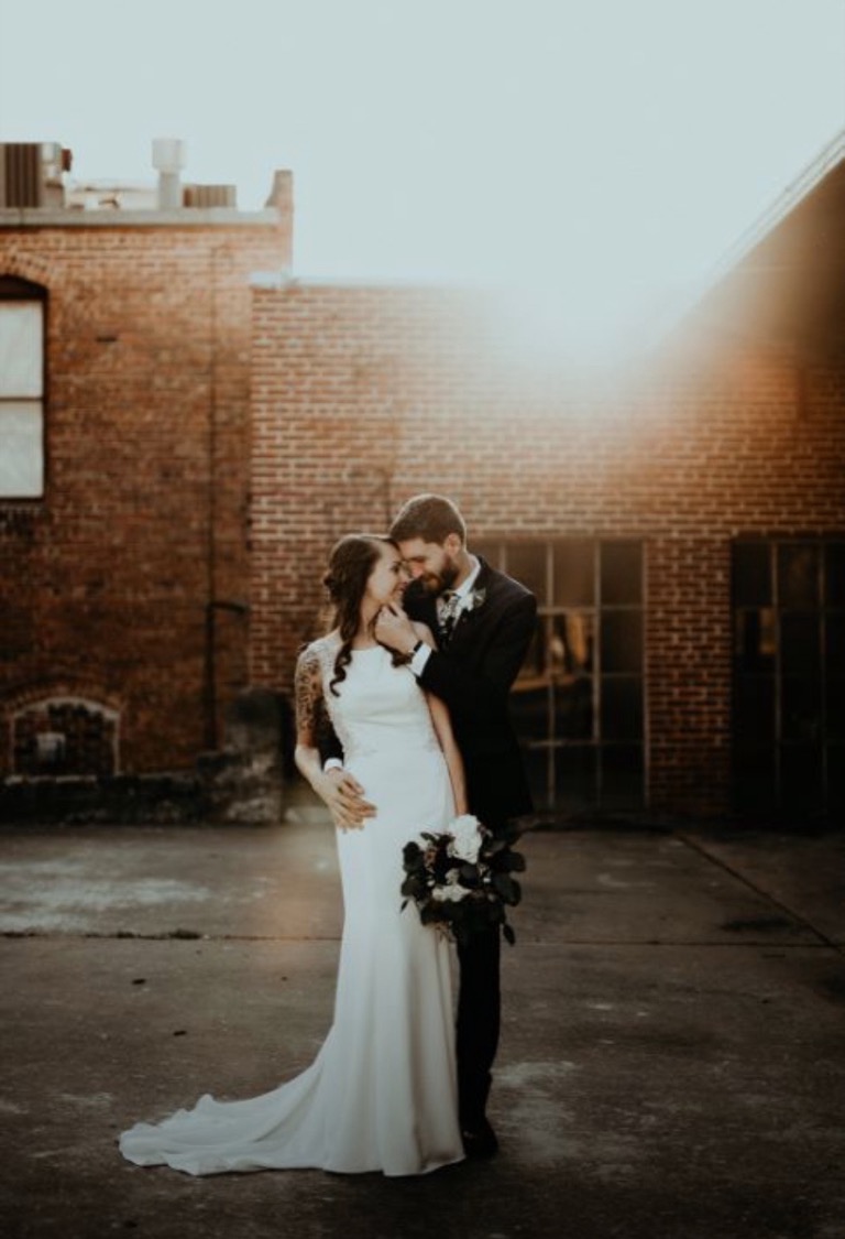 A couple against a brick wall as the sun flares low over the rooftop behind them.