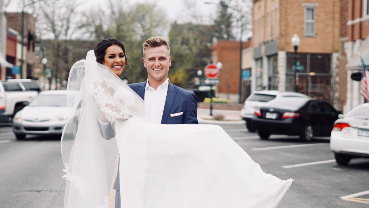 The groom carrying the bride across South Broadway, downtown storefronts and parked cars behind them.